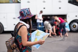 Woman with hat and headphones looking at a map near a bus with people.